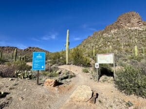 The Sarasota Trailhead entrance in Tucson Mountain Park features trail signage, a route map, towering saguaros, and a rugged Sonoran Desert mountainside in Tucson, Arizona.