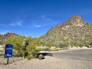 Sarasota Trailhead parking area in Tucson Mountain Park with a blue trail sign, desert landscaping, towering saguaros, and rugged mountain slopes under a vivid Arizona sky.
