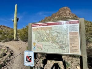 Trail map and guide sign at Sarasota Trailhead in Tucson Mountain Park, with a towering saguaro and rugged desert mountain backdrop in Tucson, Arizona.